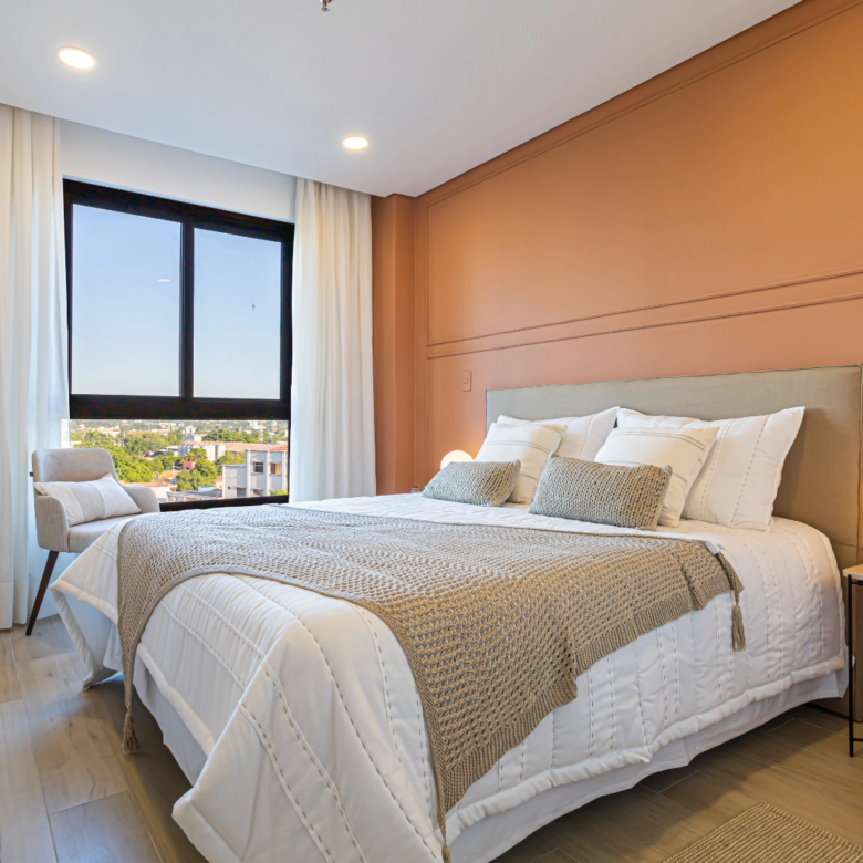 Chambre meublée avec grand lit, mur terracotta et fenêtre sur la ville dans l’immeuble Imagina Ycua Satí à Asunción, Paraguay.
